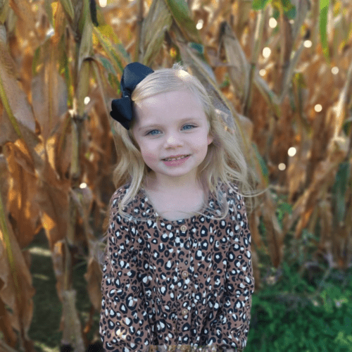 Little girl with blonde hair in a corn field with a patterned dress and black bow in her hair.